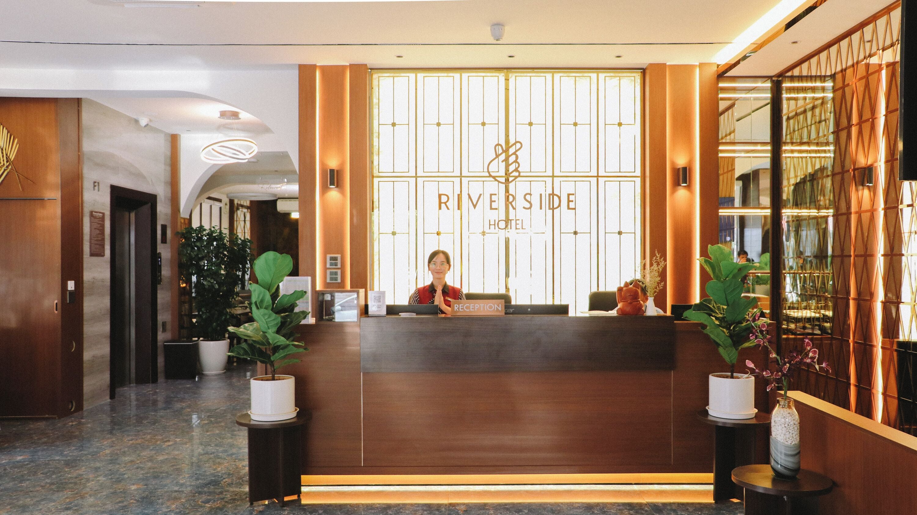 A reception desk at Riverside Hotel with a receptionist, a wooden reception desk, potted plants, and a sign that reads "RIVERSIDE HOTEL".