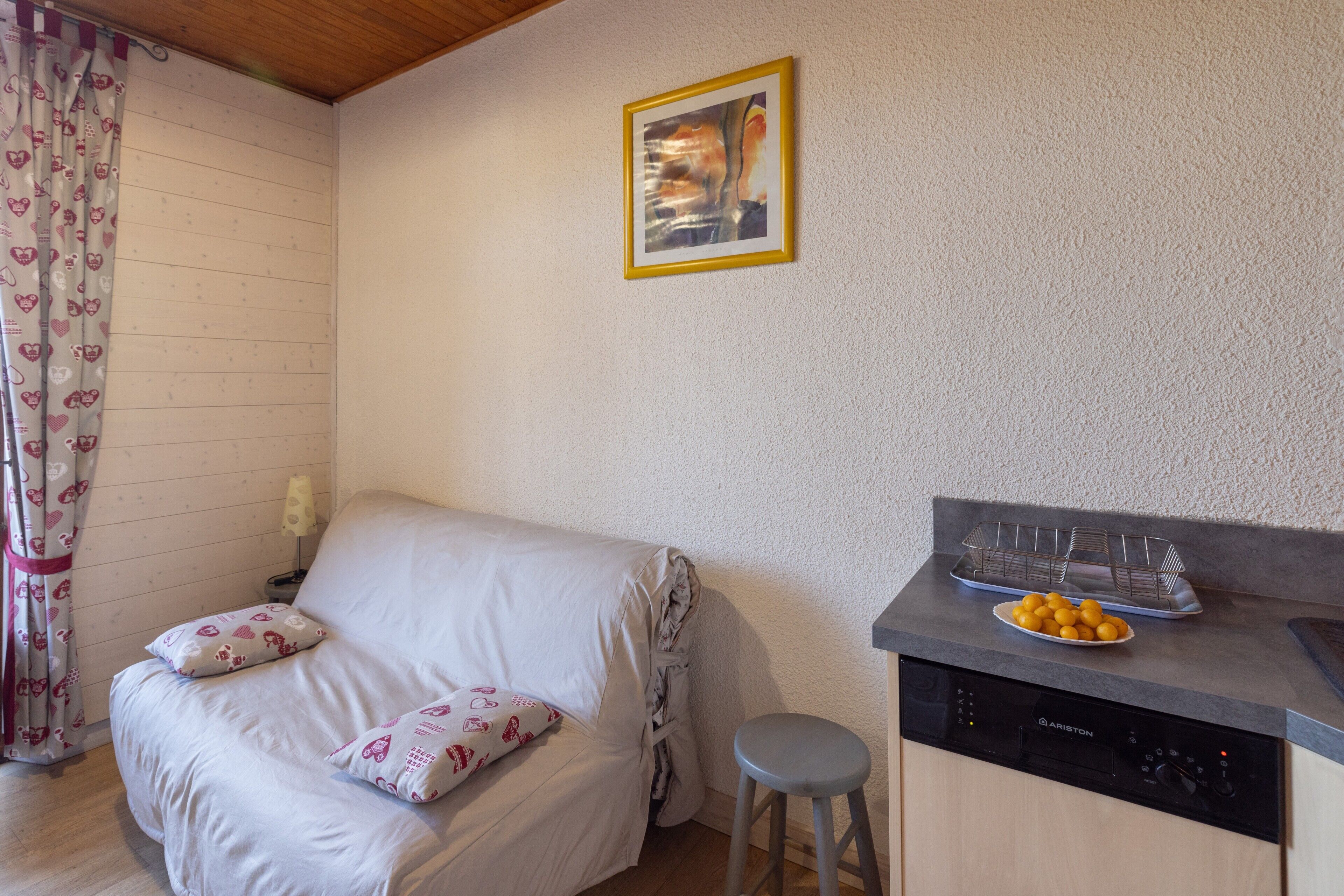 A room with a sofa, a small stool, a kitchen area with a dishwasher, and a wall-mounted picture.