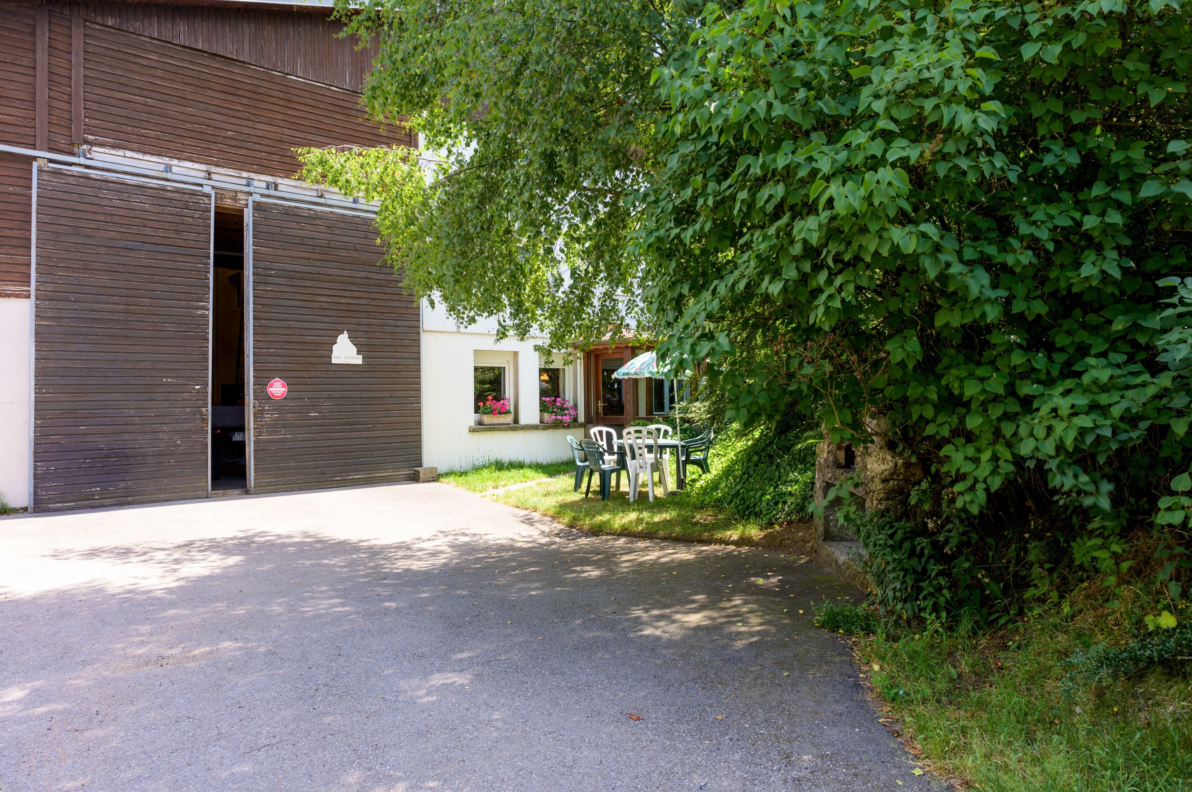 Een gebouw met een gesloten garage, een klein terras met stoelen, en een boom die schaduw geeft.