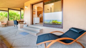 Et moderne udendørs loungeområde med stengulv, en træstol, en loungeseng med hynder og udsigt til et køkken og en balkon.