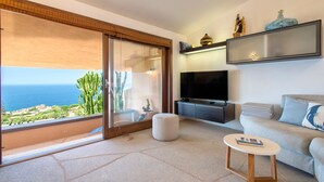Uma sala de estar moderna com uma grande porta de vidro deslizante para uma varanda, uma televisão de tela plana, um sofá, uma pequena mesa redonda e uma estante decorativa.