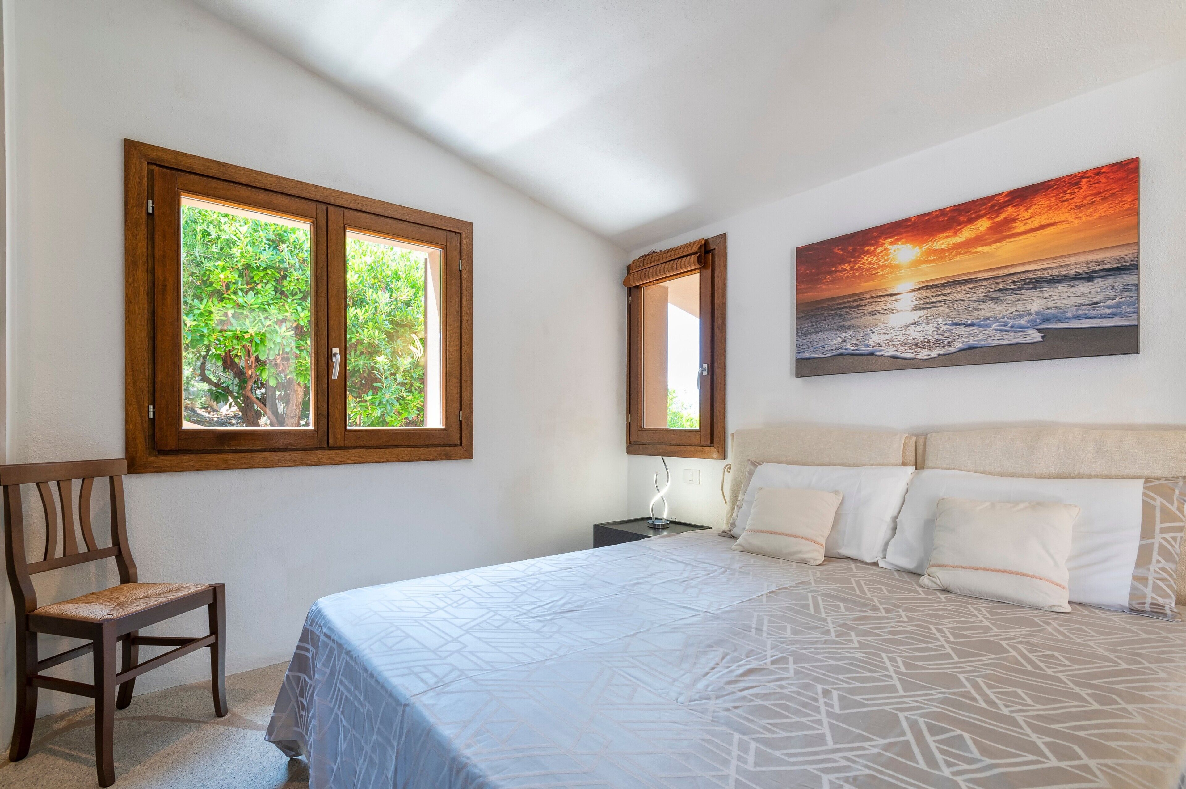 Une chambre à coucher avec un lit, deux fenêtres, une chaise et un tableau représentant un coucher de soleil.