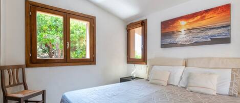 Une chambre à coucher avec un lit, deux fenêtres, une chaise et un tableau représentant un coucher de soleil.