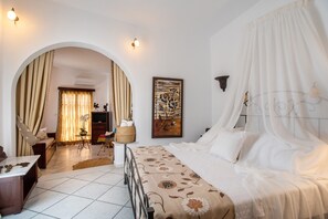 A bedroom with a bed, a desk, a chair, a small table, a plant, and a painting.