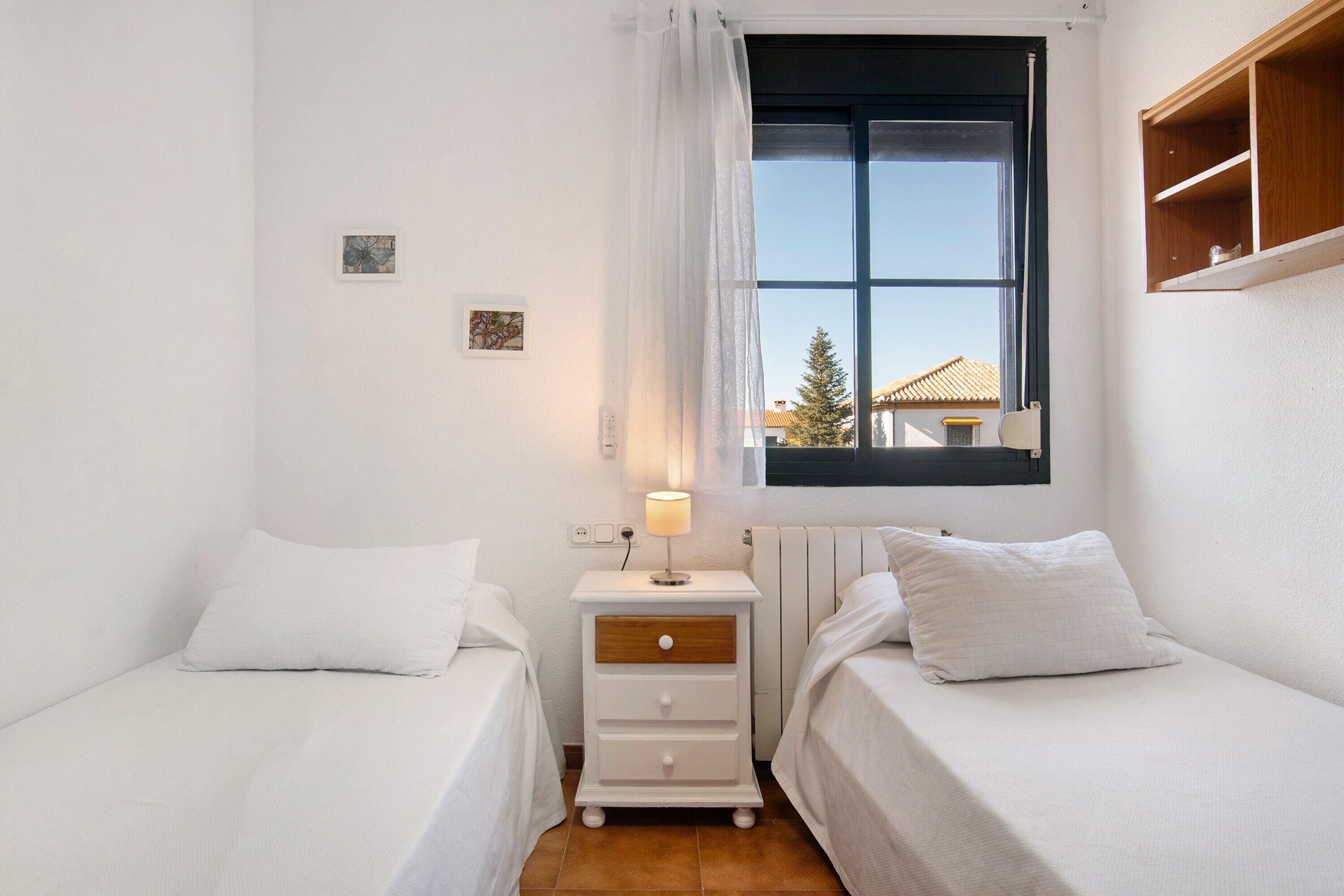 A bedroom with two beds, a nightstand with a lamp, a window with a view of a building, and a small shelf above the bed.