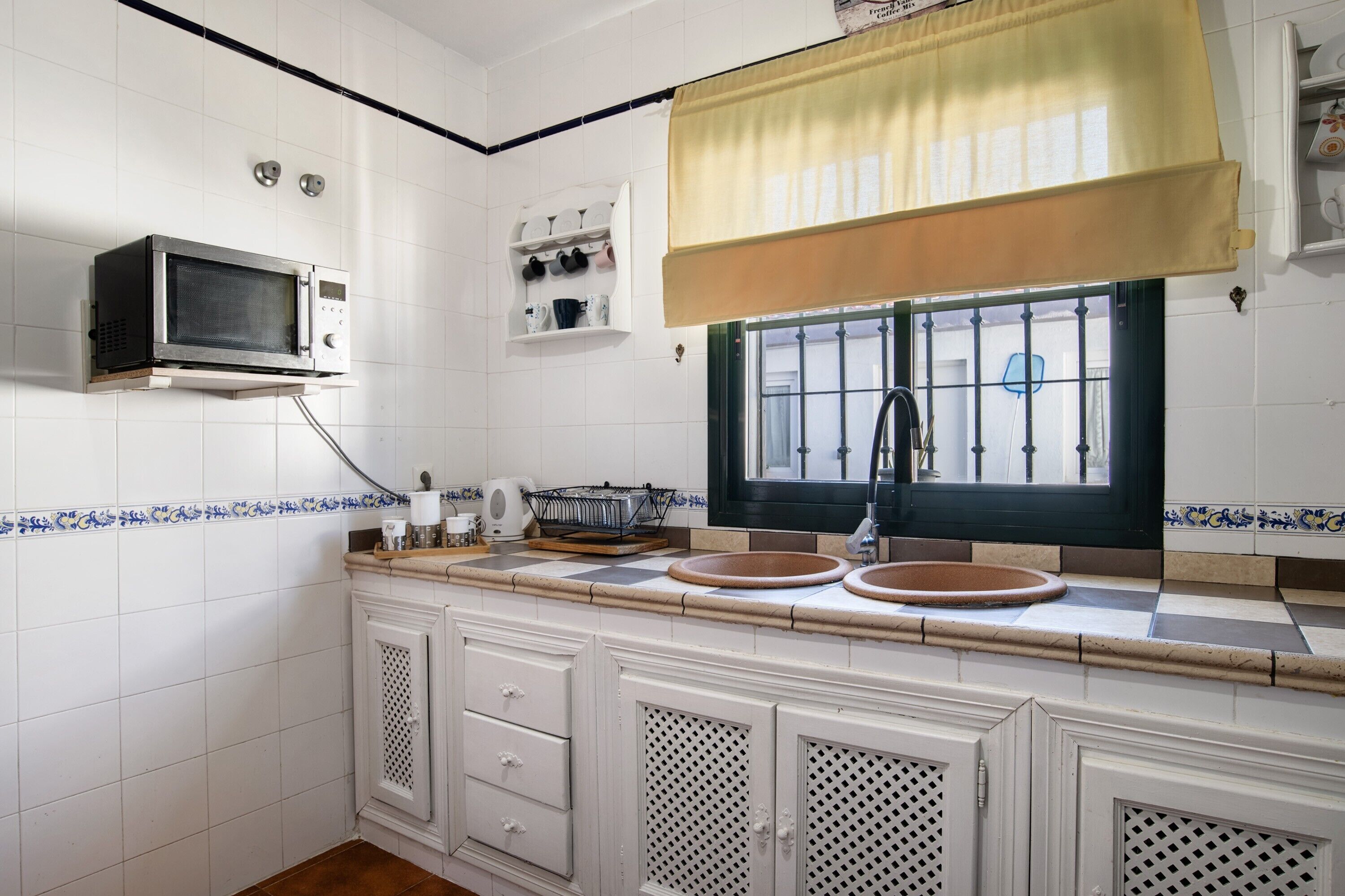 Una cocina con paredes de azulejos blancos, microondas, ventana con vistas y un lavamanos.