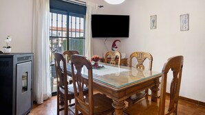 Un coin repas comprenant une table à plateau en verre, des chaises en bois, un téléviseur fixé au mur et une fenêtre avec des rideaux.