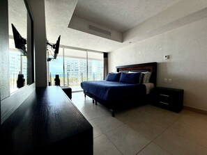 Ein Schlafzimmer mit Bett, Nachttisch, Schreibtisch und einem großen Fenster mit Blick auf Gebäude.