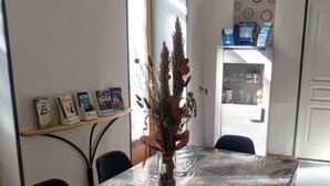 Una mesa de comedor con mantel de plástico transparente, un florero con plantas secas y una estantería con libros y un reloj.