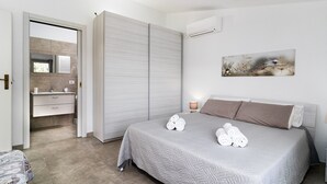 Une chambre à coucher comprenant un lit, une table de chevet, une commode, un climatiseur mural, un tableau, et une salle de bain avec un miroir, un lavabo et un porte-serviettes.