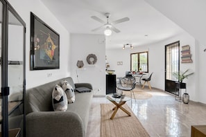 A modern living room with a grey sofa, a glass coffee table, a ceiling fan, and a dining area with chairs and a table.