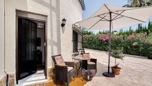 Een terras buiten met rieten stoelen, een tafel, een bloempot en een grote parasol.