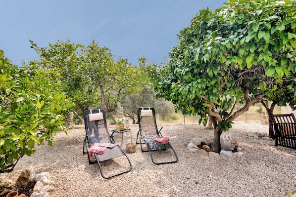 Two lounge chairs, a small table, and a statue in a garden with gravel ground and trees.