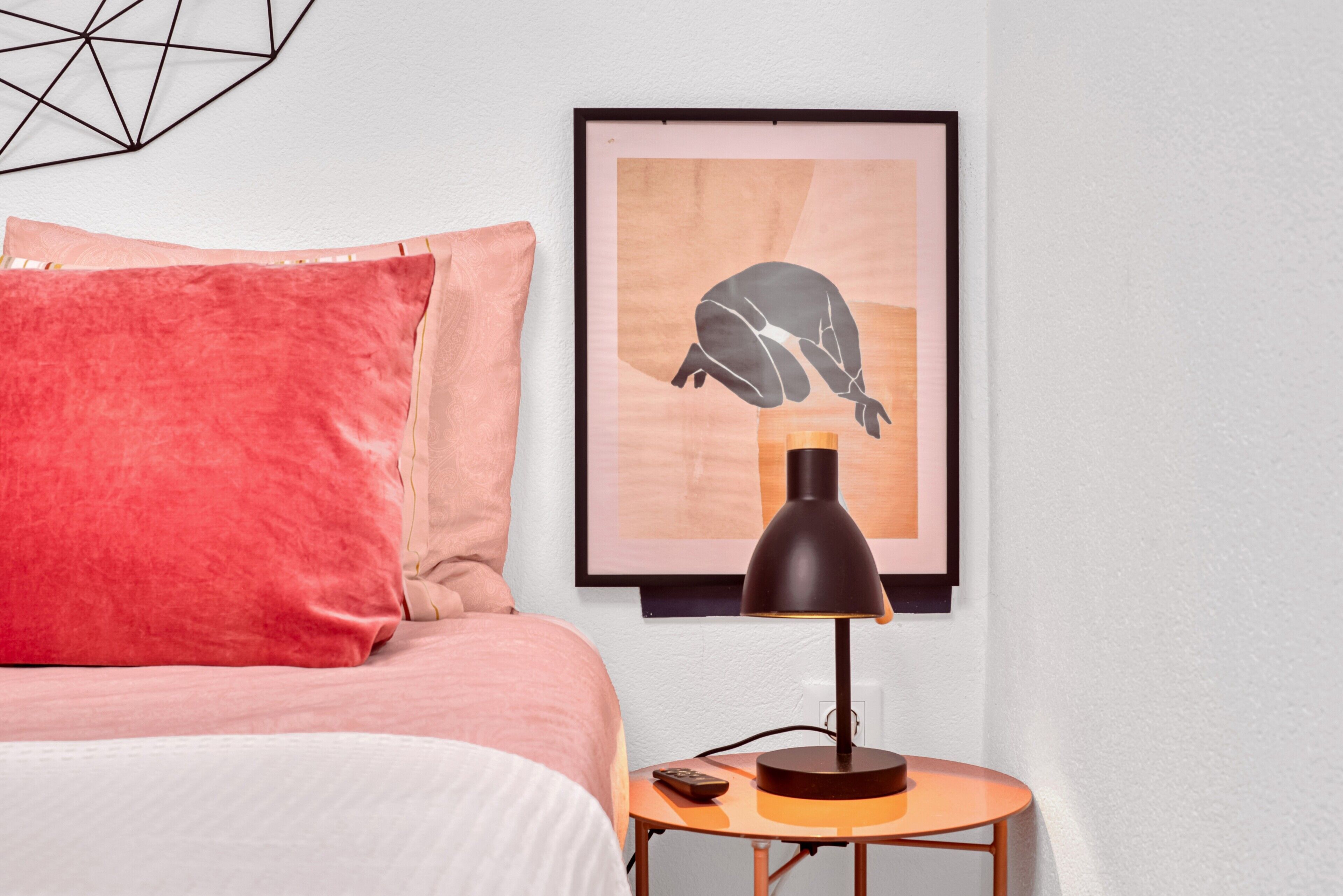 A neatly made bed with a red pillow, a framed artwork on the wall, and a small table with a lamp and remote control.