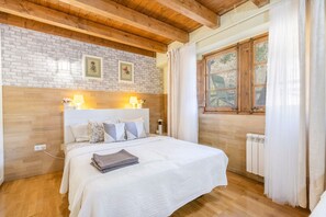 A bedroom with a wooden ceiling, a brick wall, a large window with curtains, a bed with white bedding, and two wall-mounted lamps.