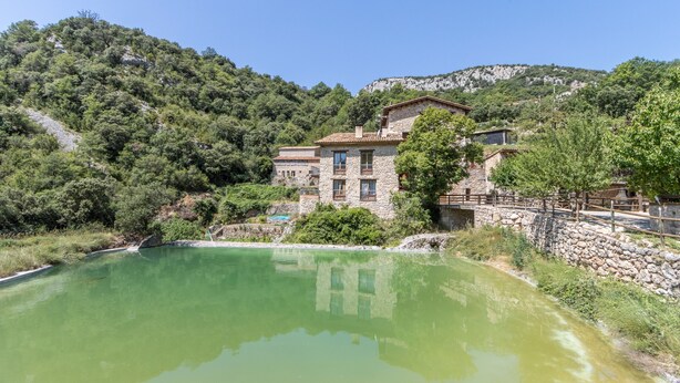 Un bâtiment en pierre doté d’un balcon, entouré de verdure et bordé par un plan d’eau.