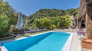 Une piscine entourée de verdure et bordée d’une colline, avec des chaises longues et des parasols.