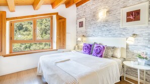 Une chambre avec une grande fenêtre, un lit recouvert d’une courtepointe à rayures, un mur en pierre et une petite table de chevet avec une lampe.