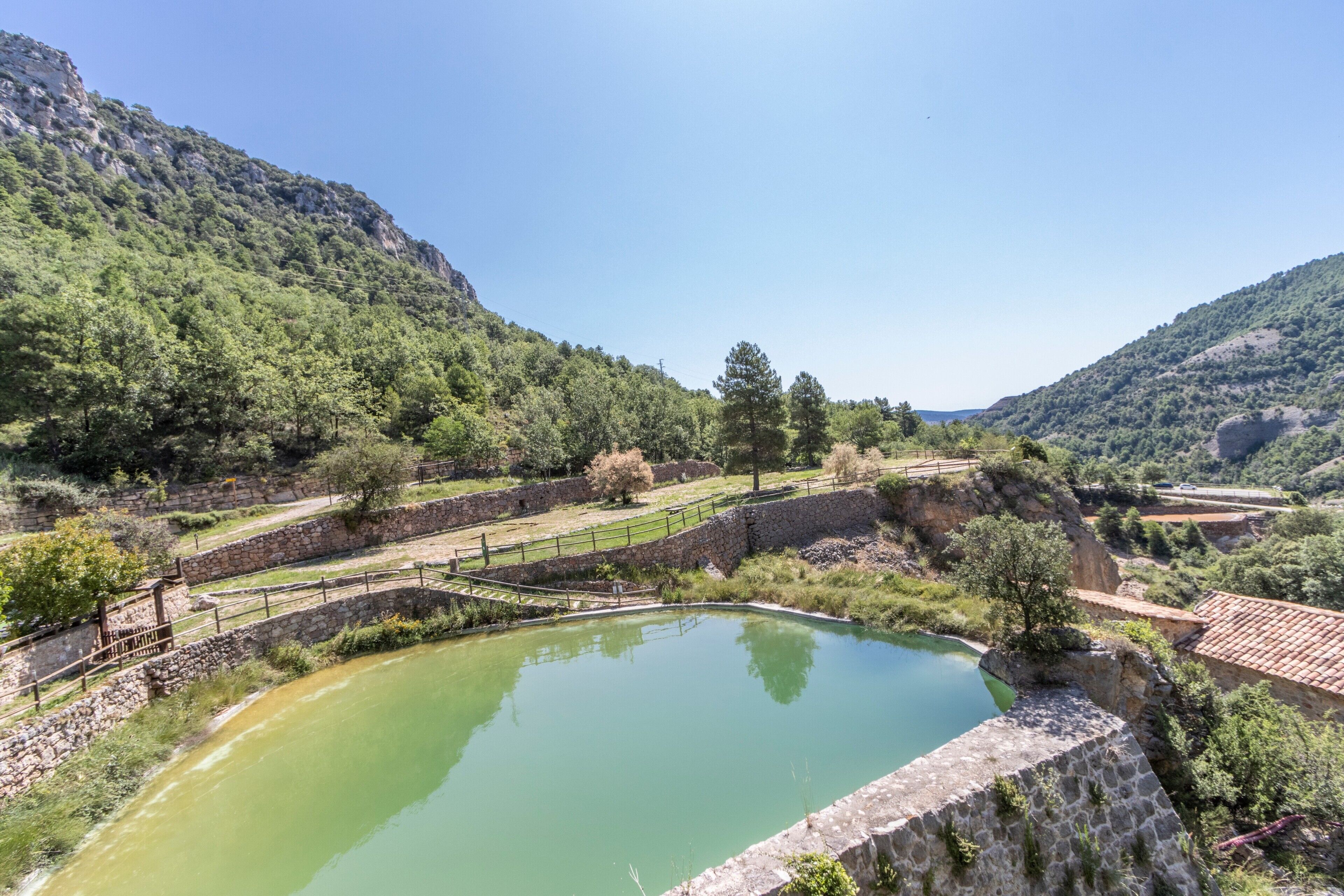 Paisatge natural amb una bassa, parets de pedra i una vessant boscosa.