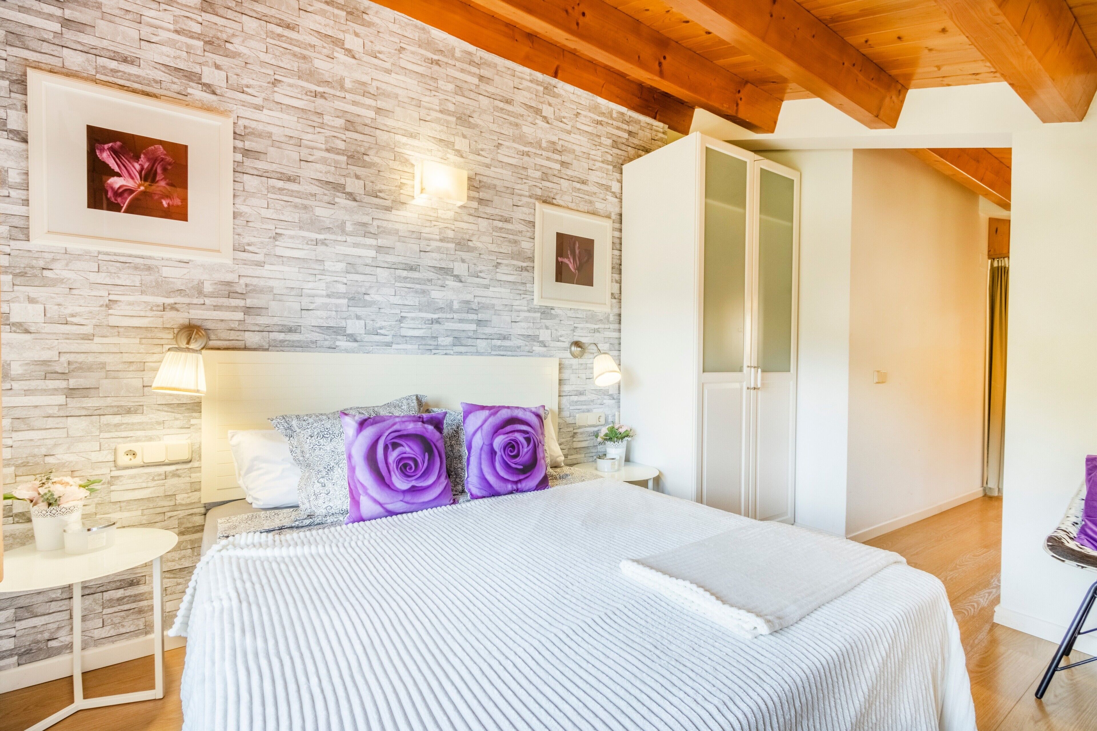 Quarto com parede de pedra, cama com colcha listrada em branco e azul, almofadas de rosas roxas, mesa de cabeceira branca com abajur e teto de madeira.