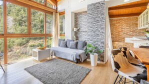 A modern bedroom with a bed, a stone accent wall, a dining area with a wooden table and chairs, and large windows offering a view of the outdoors.