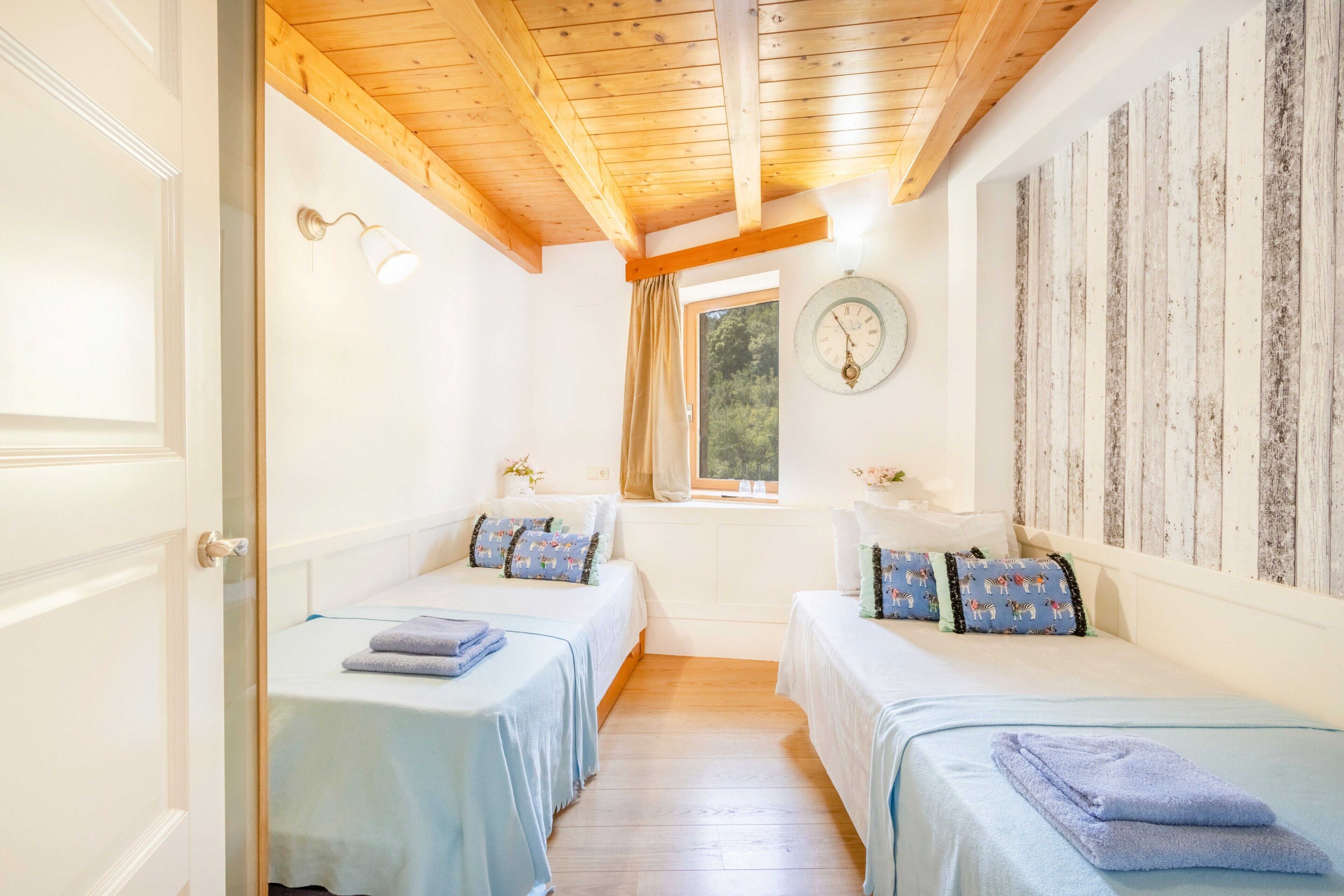 A room with two beds, a wooden ceiling, a clock on the wall, and a window with curtains.
