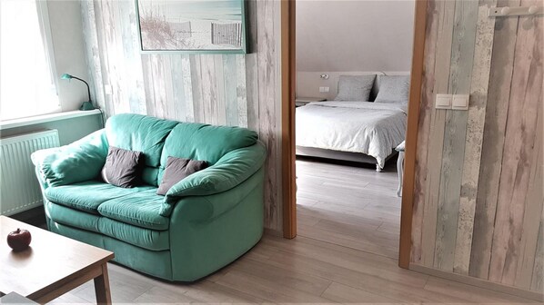 Una habitación con un sofá azul verdoso, piso de madera, cama con colcha gris y pared de madera con un cuadro.
