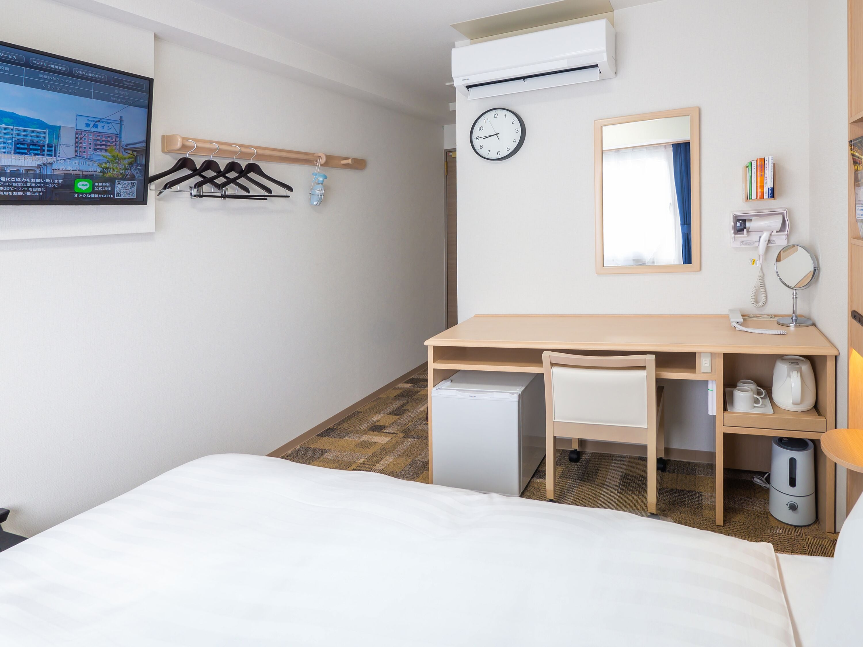 Hotellihuone, jossa on sänky, työpöytä tuolilla, pieni jääkaappi, seinälle asennettu televisio, kello, peili, hylly hygieniatuotteille ja naulakko.