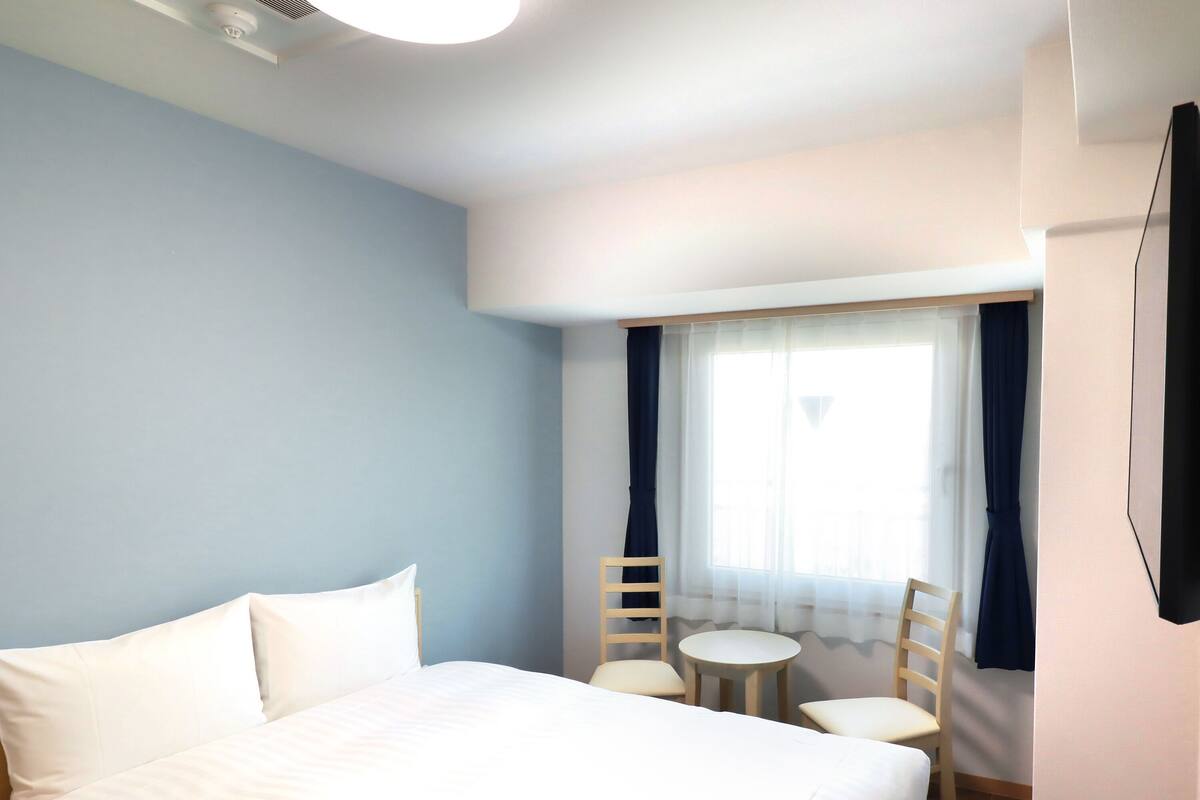 Habitación de hotel con una cama grande, una mesita redonda pequeña, dos sillas, una ventana con cortinas y un televisor de pantalla plana montado en la pared.