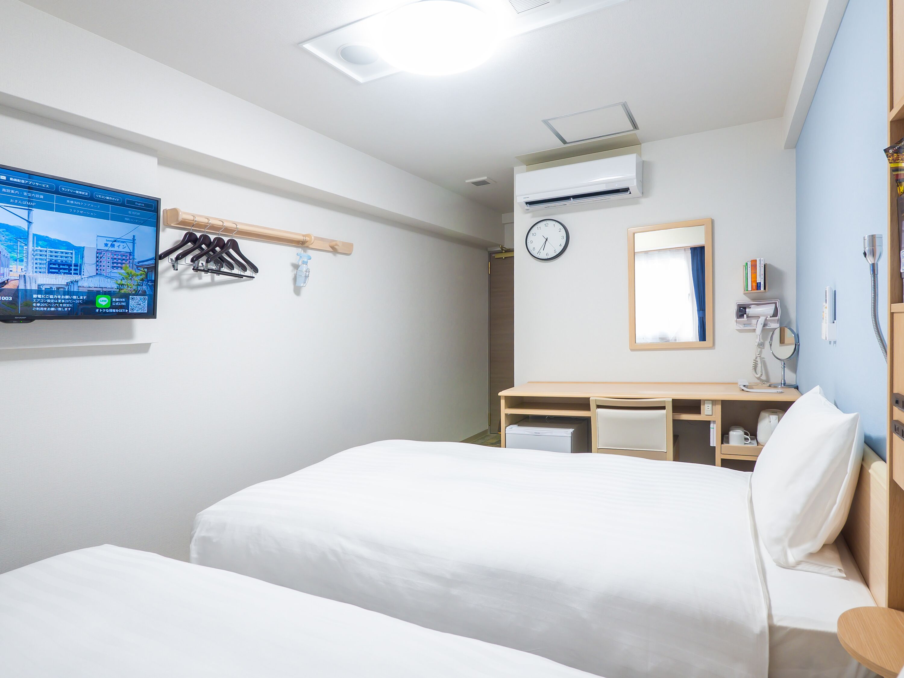 A hotel room with two beds, a desk, a clock, a mirror, a TV, and a window.