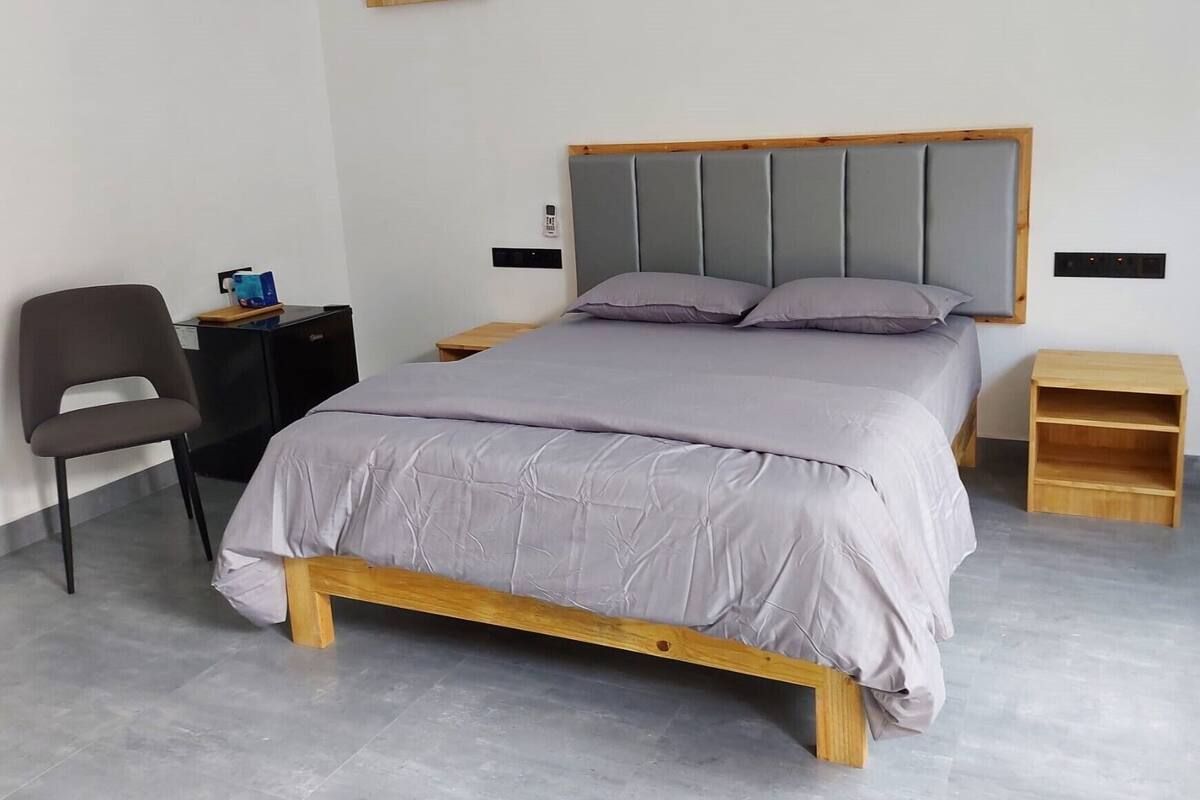 A modern bedroom with a wooden bed frame, grey bedding, a grey upholstered headboard, a wooden nightstand, a small wooden shelf, and a grey office chair.