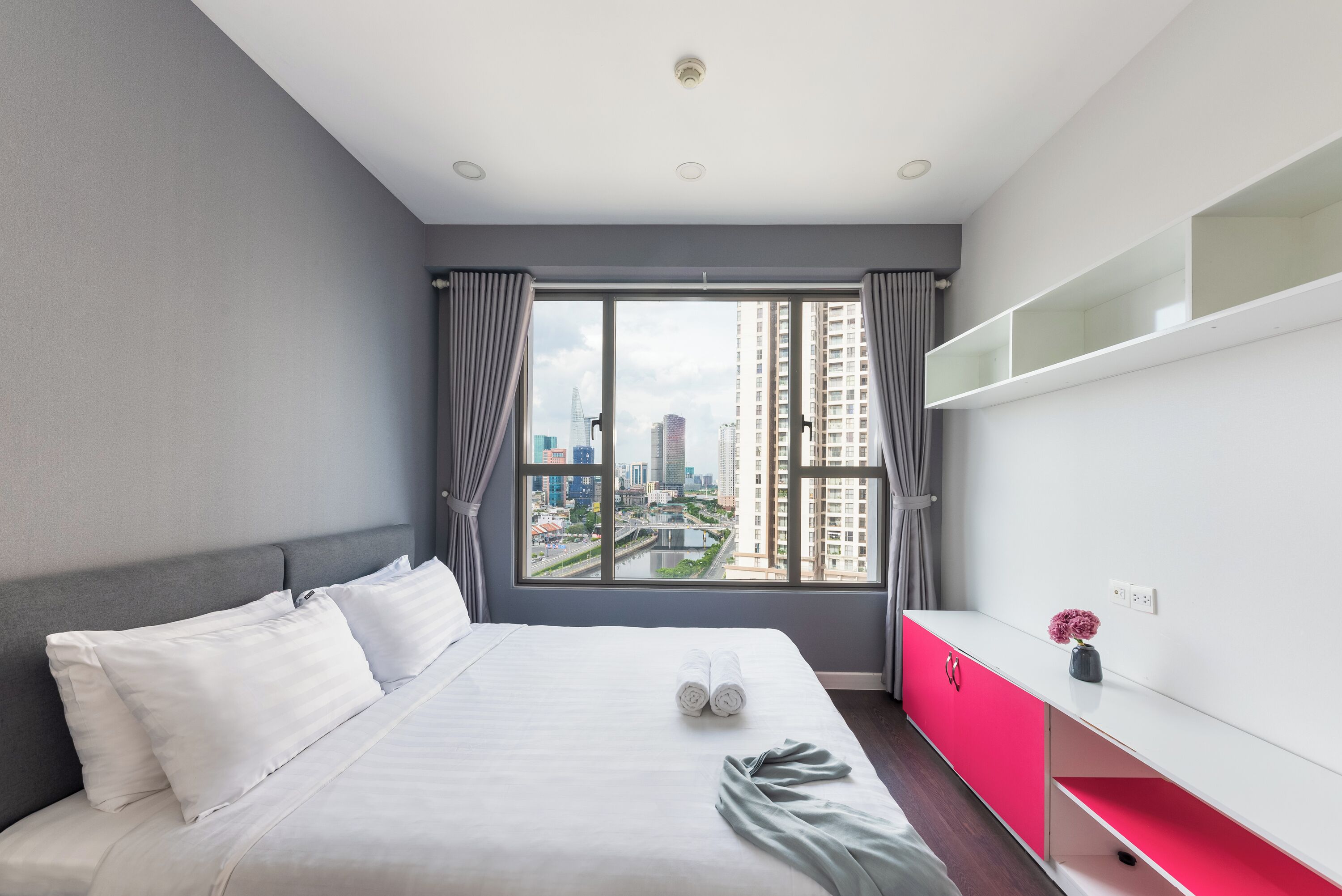 A modern hotel room with a large bed, a city view, a white console table, and a red cabinet.