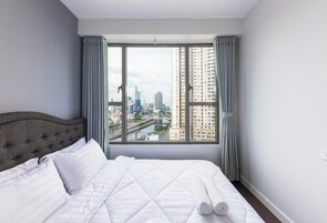 Habitación de hotel con una cama grande, cortinas grises y vista a un paisaje urbano a través de un ventanal amplio.