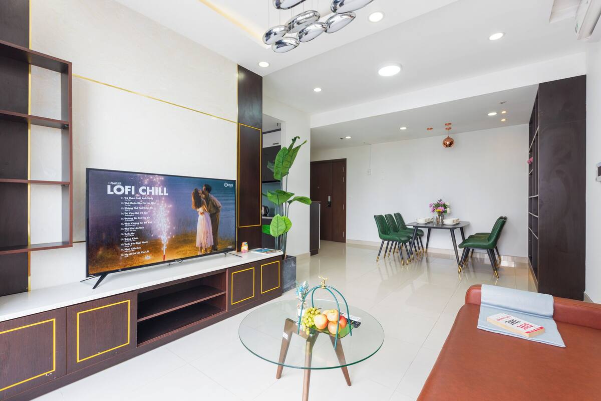 A modern living room with a flat-screen TV, a glass coffee table with fruit, a sofa with a book, and a dining area with green chairs.