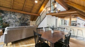 A spacious loft with a stone wall, wooden ceiling, a dining area with a table and chairs, a sofa, and a kitchen in the background.