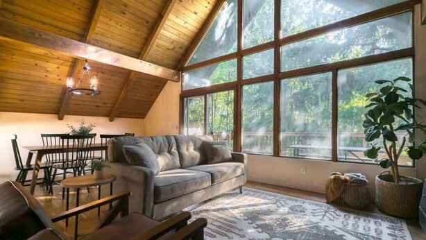 A cozy room with a wooden ceiling, large windows, a sofa, a dining table, and chairs.