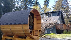 A wooden barrel-shaped cabin with a sloped roof, a small porch, and a view of a house in the background.