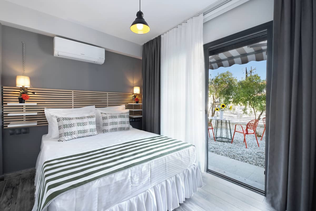 Ein modernes Hotelzimmer mit einem großen Bett, Nachttischlampen und Blick auf eine Terrasse mit roten Stühlen und Sonnenblumen.