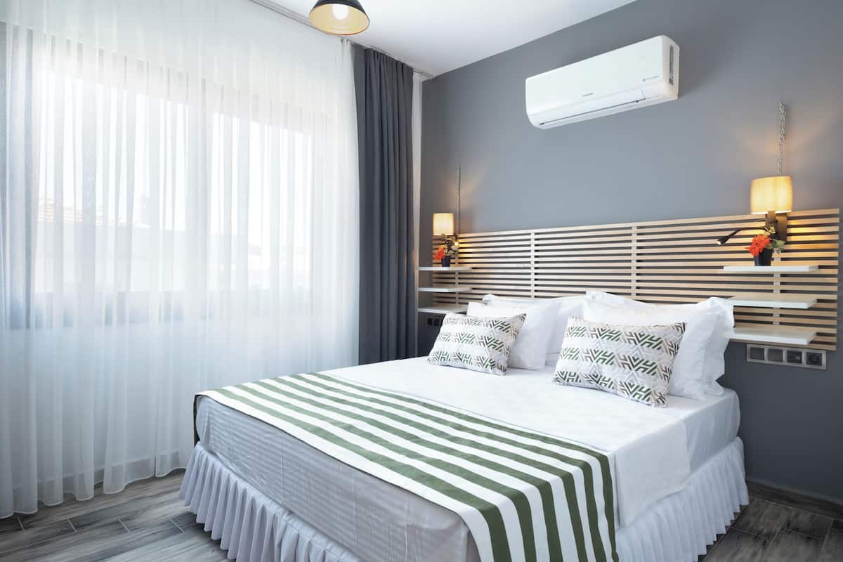 Ein modernes Hotelzimmer mit einem großen Bett, einem Fenster mit durchscheinenden Vorhängen, einer Wand-Klimaanlage und einem Nachttisch mit Lampe.