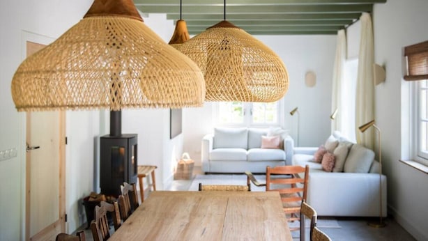 Un comedor con una mesa y sillas de madera, una lámpara colgante tejida y un sofá al fondo.