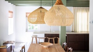 Un comedor con una mesa de madera, dos lámparas colgantes tejidas y vistas a una cocina al fondo.