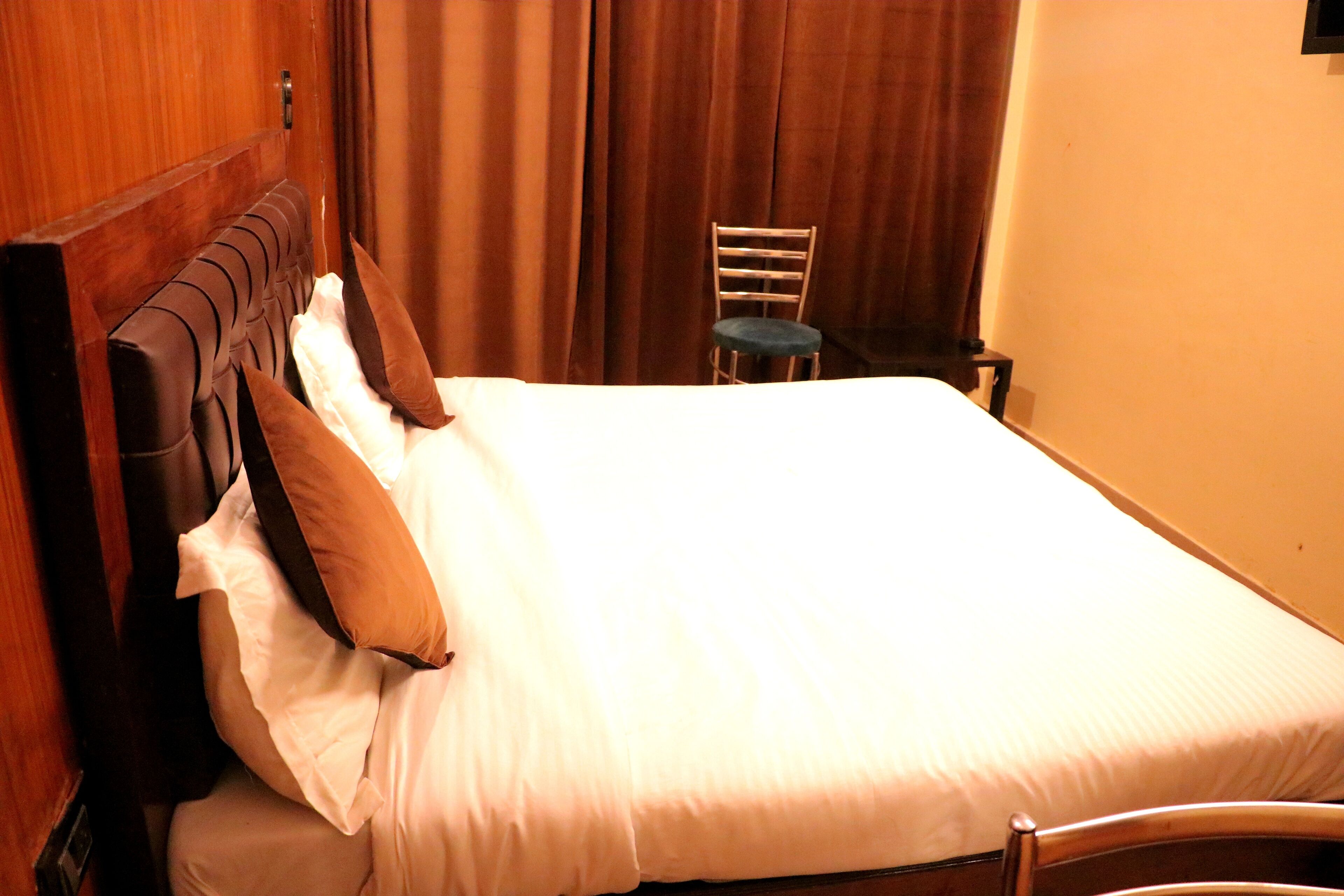 A neatly made bed with white linens and brown pillows, a wooden headboard, and a chair against a wall with curtains.