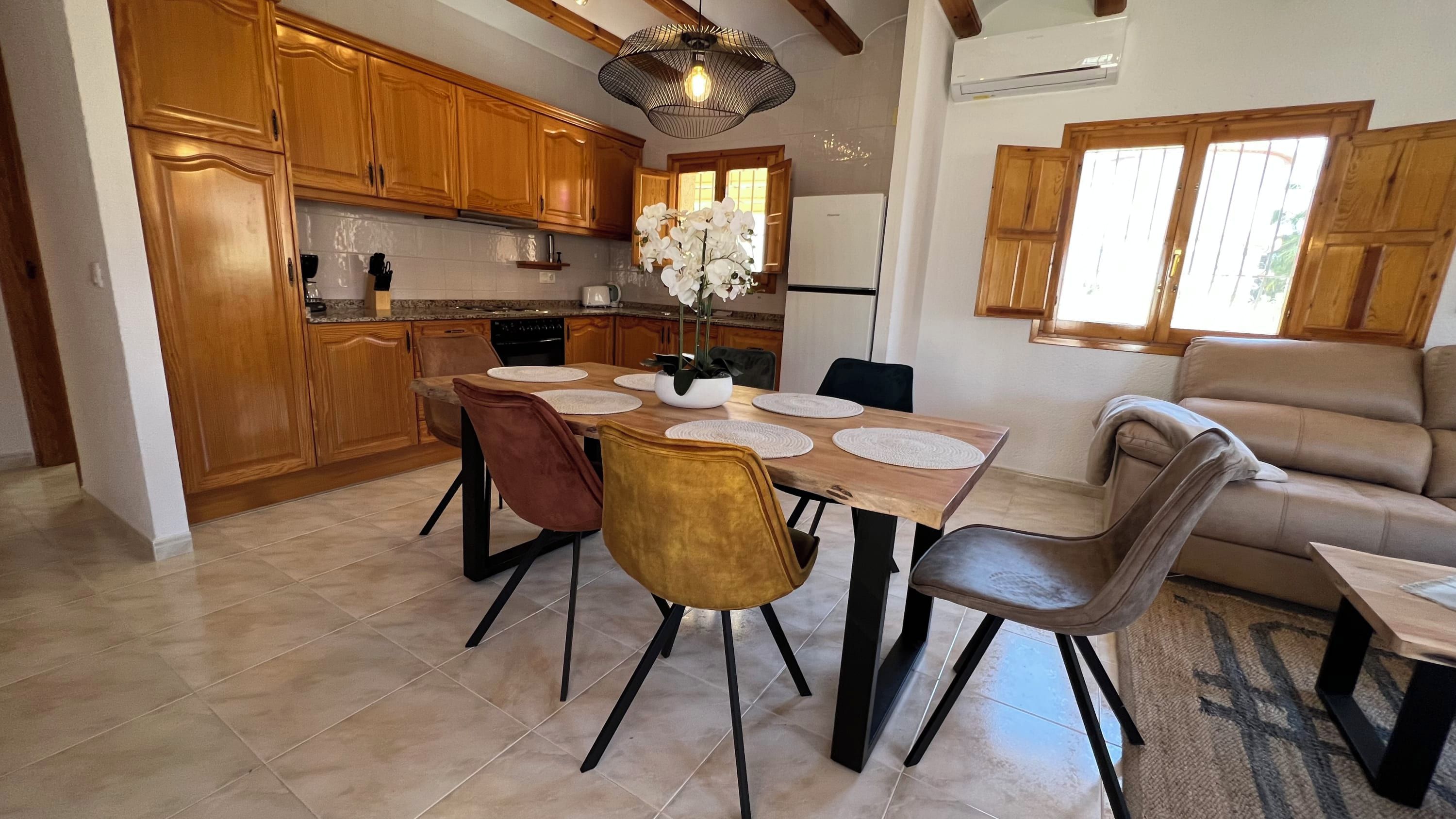 A modern kitchen with wooden cabinets, a dining table with chairs, and a living area with a sofa.