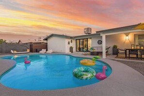 Une piscine agrémentée de flamants roses gonflables, un patio couvert et une maison moderne dotée d’un toit plat.