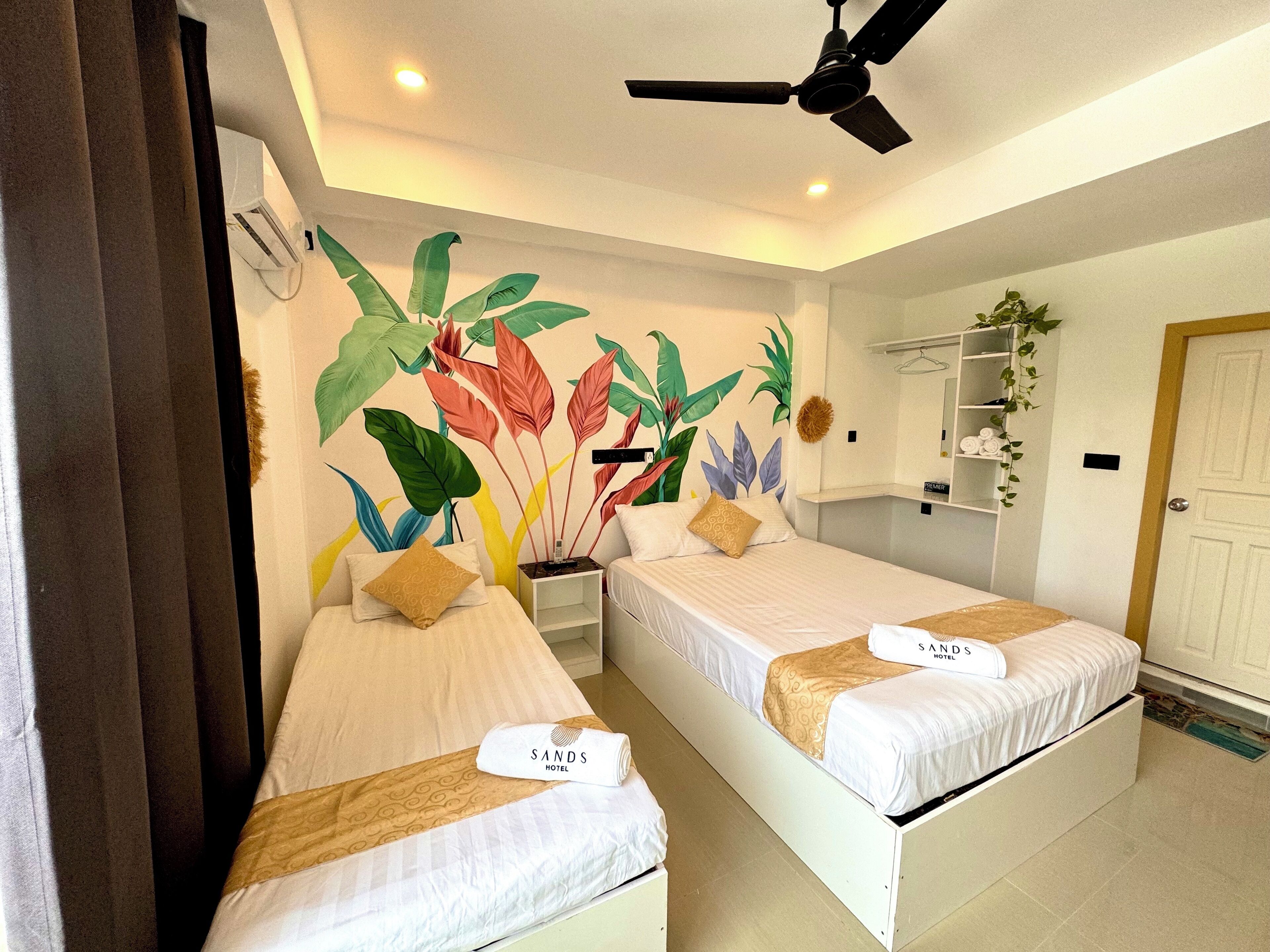 Habitación de hotel con dos camas, un ventilador de techo, estante con toallas y un mural con motivos tropicales.
