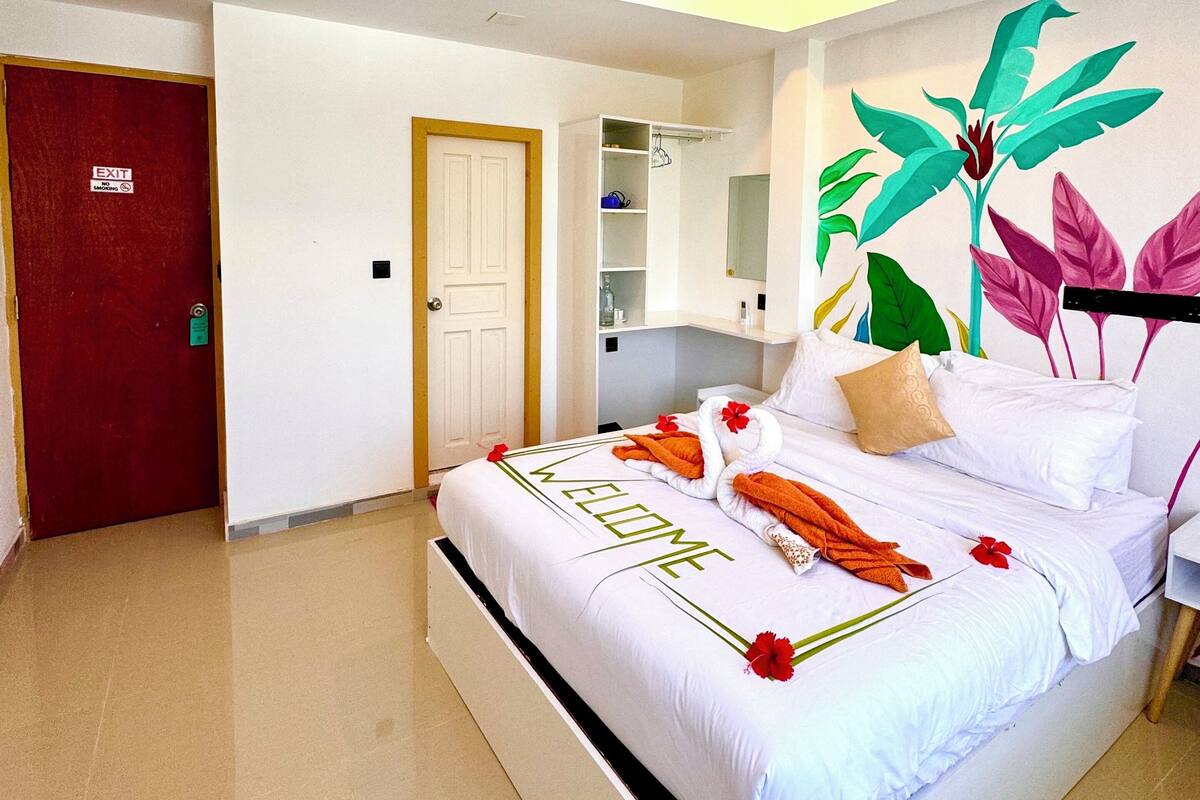 Una camera d'albergo con un letto, una ventola a soffitto, una porta rossa e un murale con piante tropicali sulla parete.