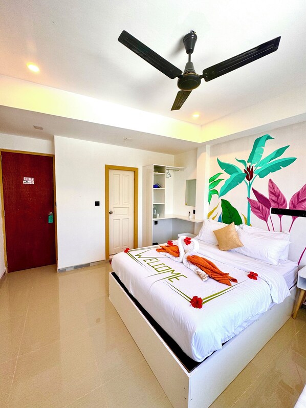A hotel room with a bed, a ceiling fan, a red door, and a wall with a tropical plant mural.