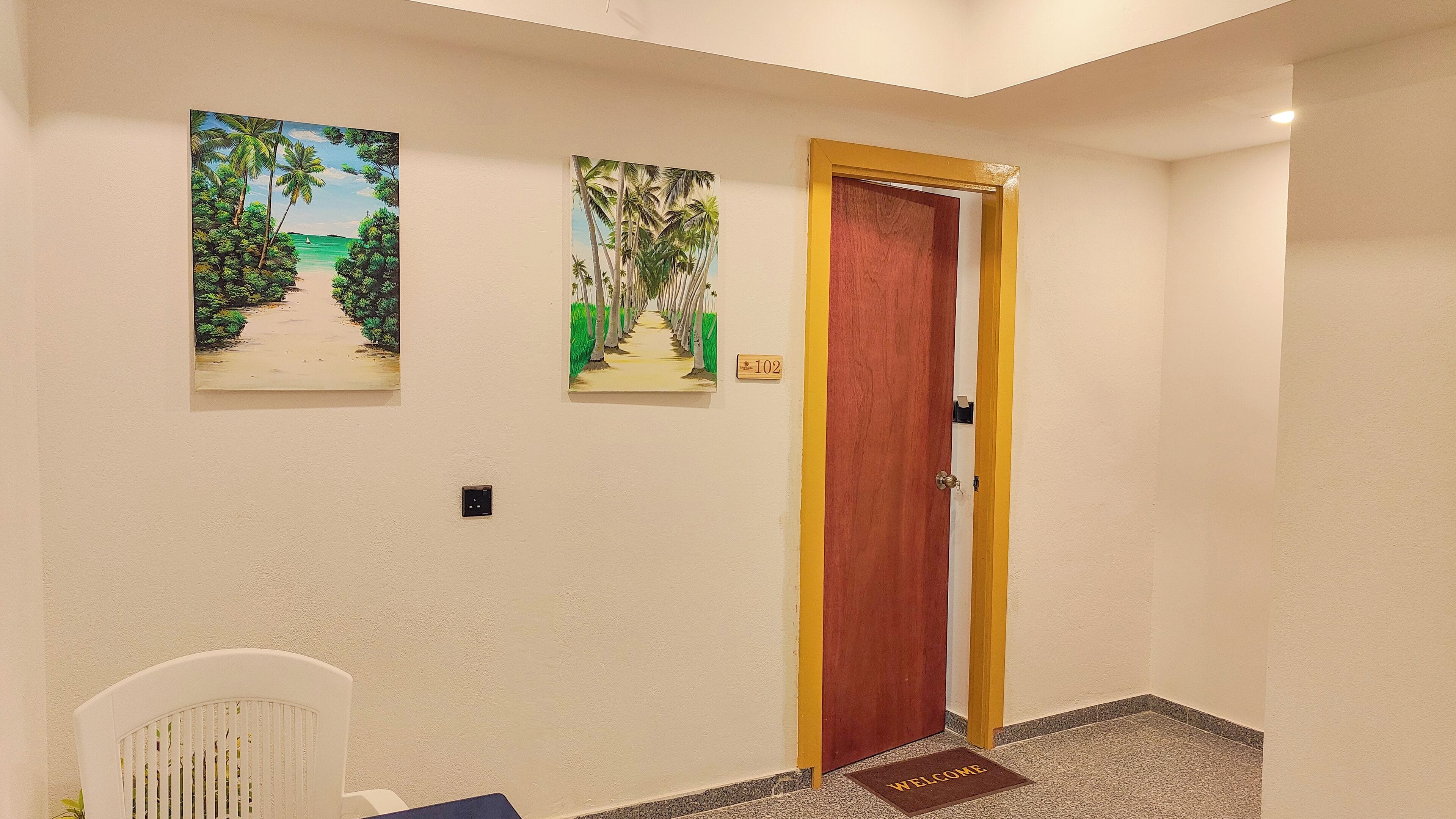 A hallway with two paintings of tropical scenes, a wooden door with a yellow frame, and a welcome mat on the floor.