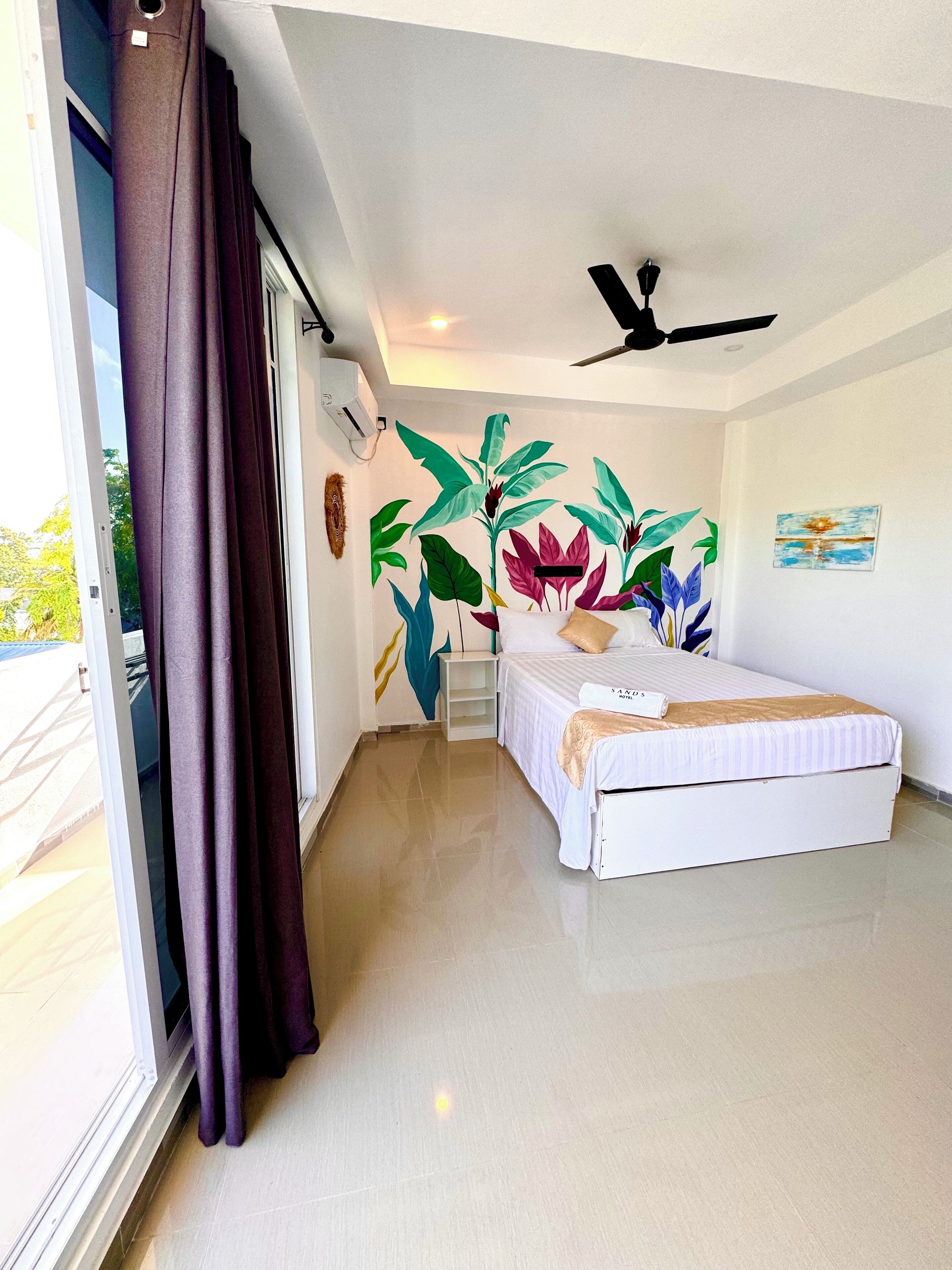 A hotel room with a bed, a ceiling fan, a window with curtains, and a wall with a tropical plant mural.
