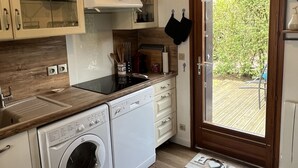 Une cuisine compacte équipée d’un lave-linge, d’une cuisinière et d’un évier. On y trouve un plan de travail en bois et une porte donnant sur un espace extérieur.